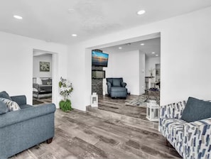 Un salon moderne avec un canapé gris, un fauteuil bleu et un canapé à motifs. Un téléviseur à écran plat est fixé au mur, et le sol est en bois.