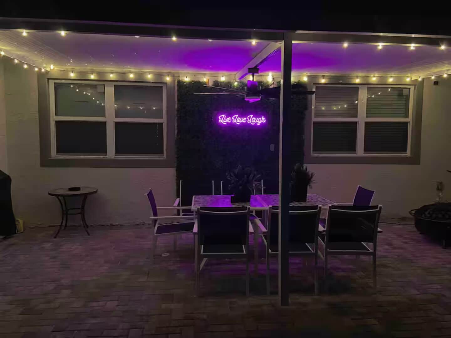 Un patio al aire libre con un letrero de neón, una mesa con sillas y guirnaldas luminosas.