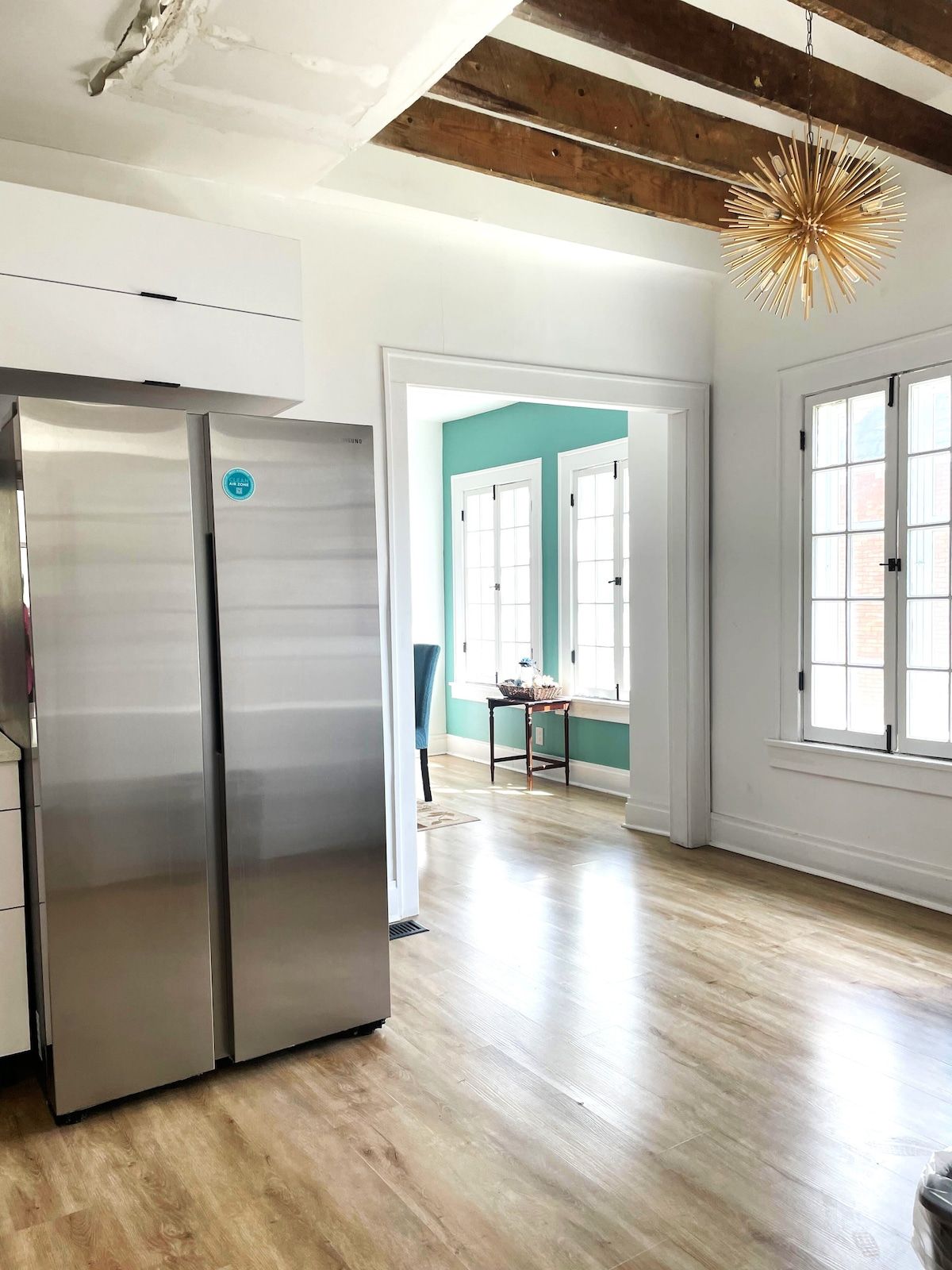 Una cocina con refrigerador de acero inoxidable, vigas de madera en el techo y vista a otra habitación con paredes azul verdosas.