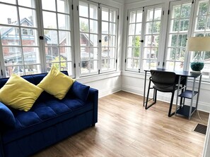 Une pièce avec un canapé bleu, des coussins jaunes, une table à manger avec une chaise, et de grandes fenêtres.