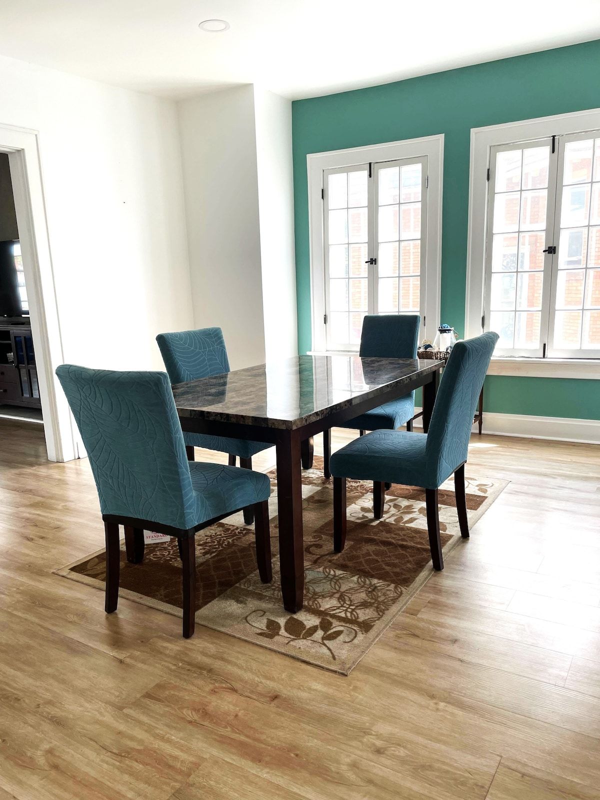 A dining area with a glass-top table surrounded by four chairs, a patterned rug, and a view of a brick building through large windows.