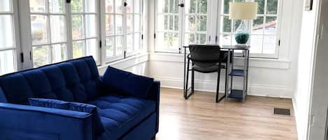 A room with a blue sofa, a chair, a small table with a lamp, and large windows.