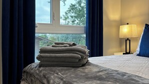 Ein Hotelzimmer mit Bett, einem Fenster mit Vorhängen, einem Stapel Handtüchern und einer Nachttischlampe.