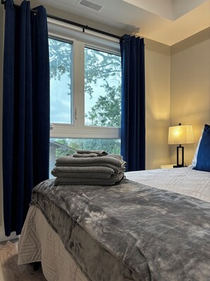 A hotel room with a bed, a window with curtains, a stack of towels, and a bedside lamp. - 1 Bedroom Condo/6 minutes from Niagara Falls. (Niagara Falls)