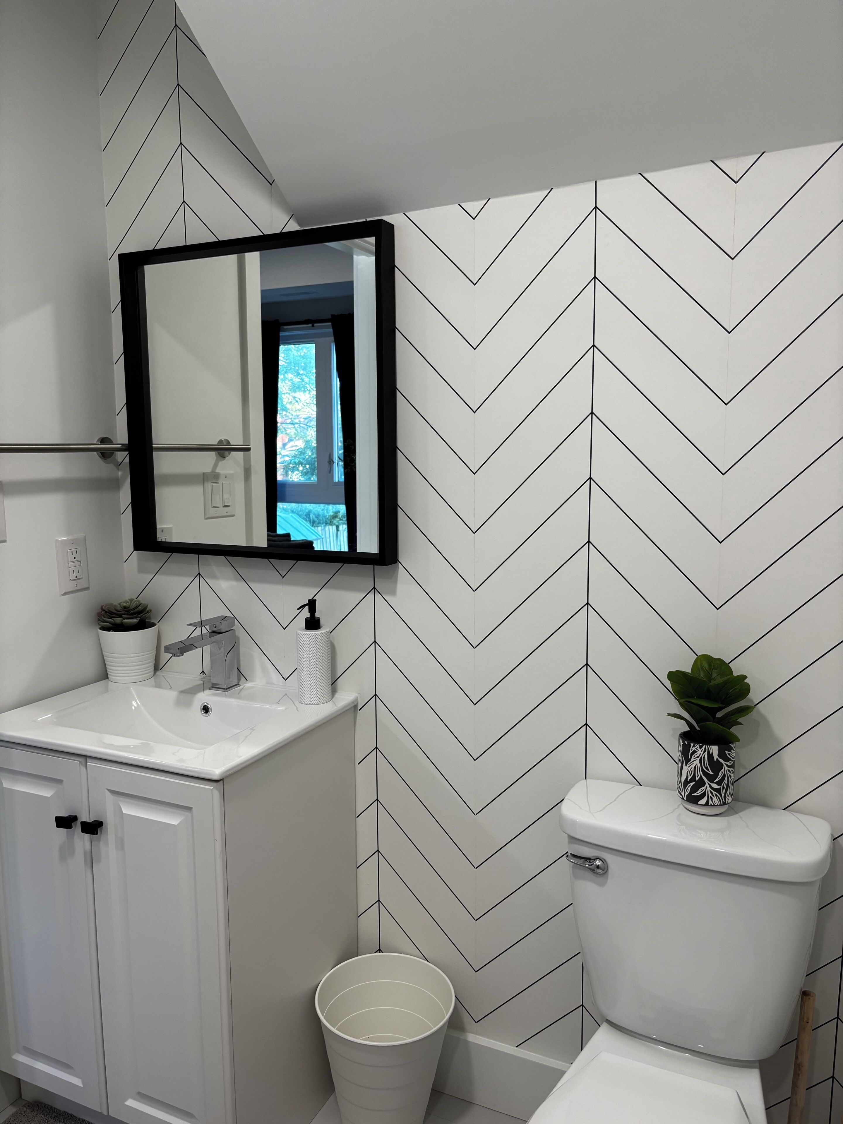 A bathroom with a white sink, mirror, and toilet. There is a white bucket and a potted plant on the toilet.
