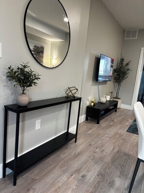 A modern living room with a black console table, a round mirror, a flat-screen TV, and a potted plant. - 1 Bedroom Condo/6 minutes from Niagara Falls. (Niagara Falls)