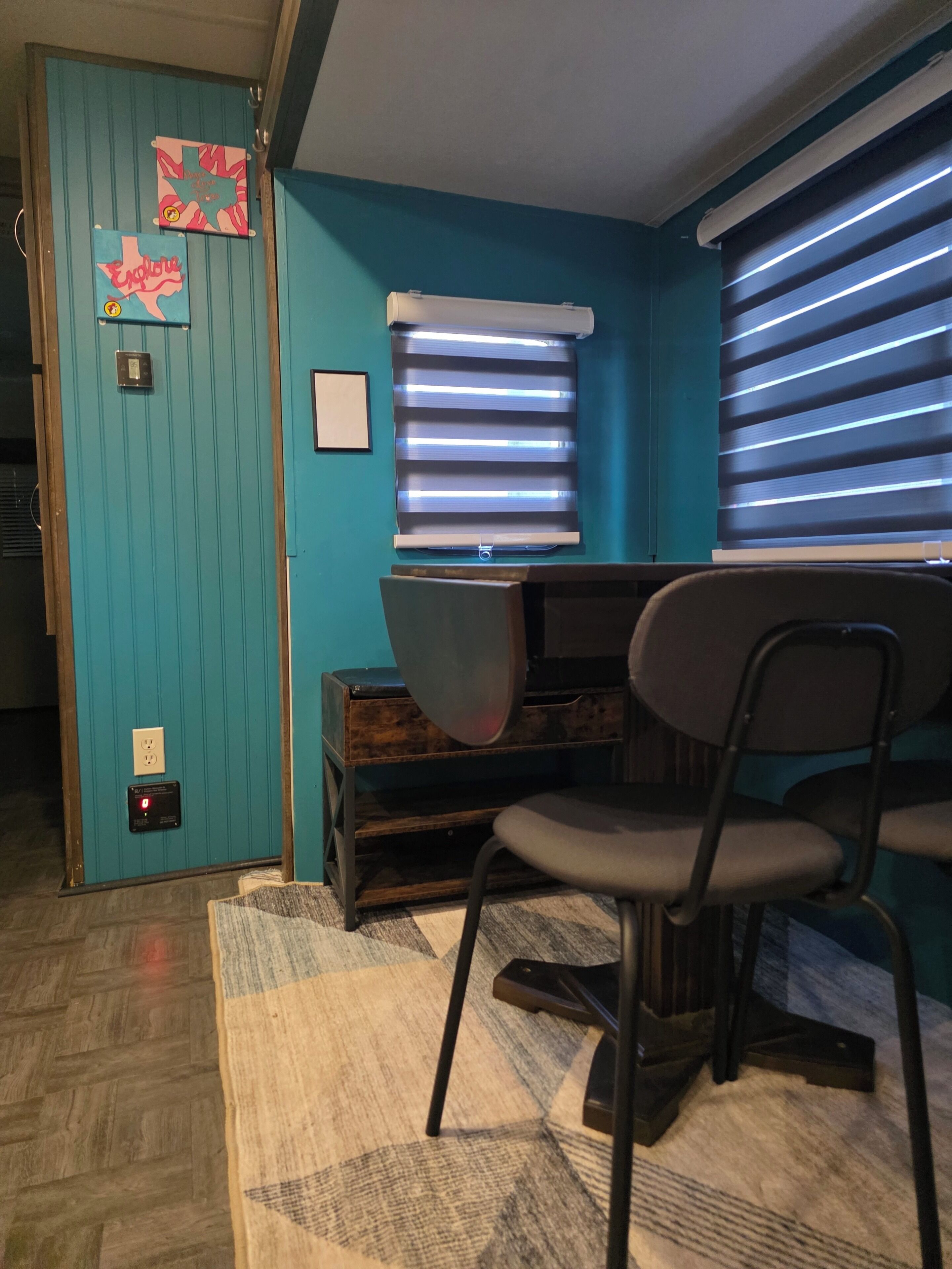 A room with teal walls, a desk with a chair, a window with blinds, and a rug on the floor.