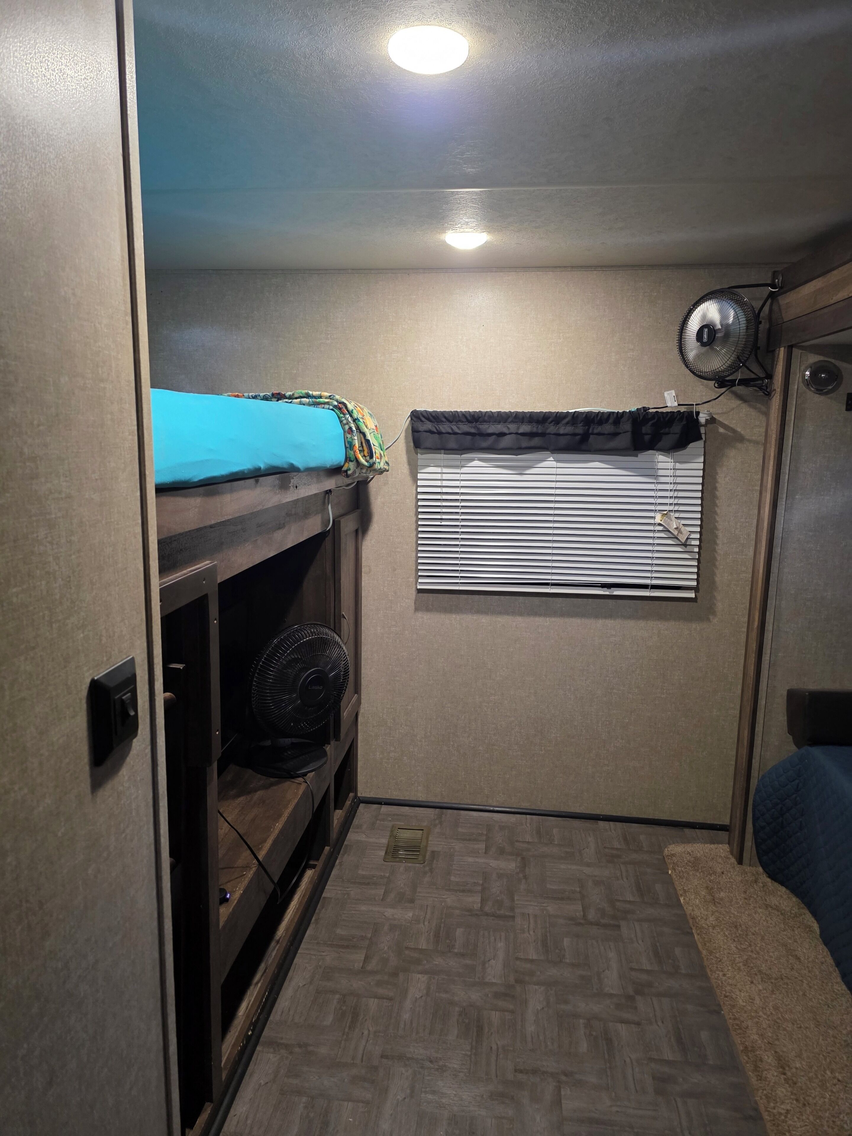 A compact room with a bunk bed, a window with blinds, a wall-mounted fan, and a ceiling light.