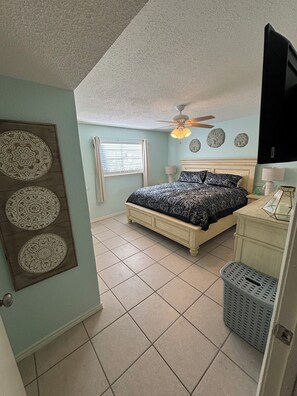 A bedroom with a bed, a ceiling fan, a window with blinds, a wall-mounted TV, and decorative plates. - Bayside Breeze: King Bed • Beach Gear Included • Fast WiFi • Pet Friendly (Galveston)