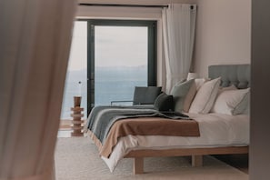 Ein Schlafzimmer mit Bett, Nachttisch, Lampe und Meerblick durch ein Fenster.