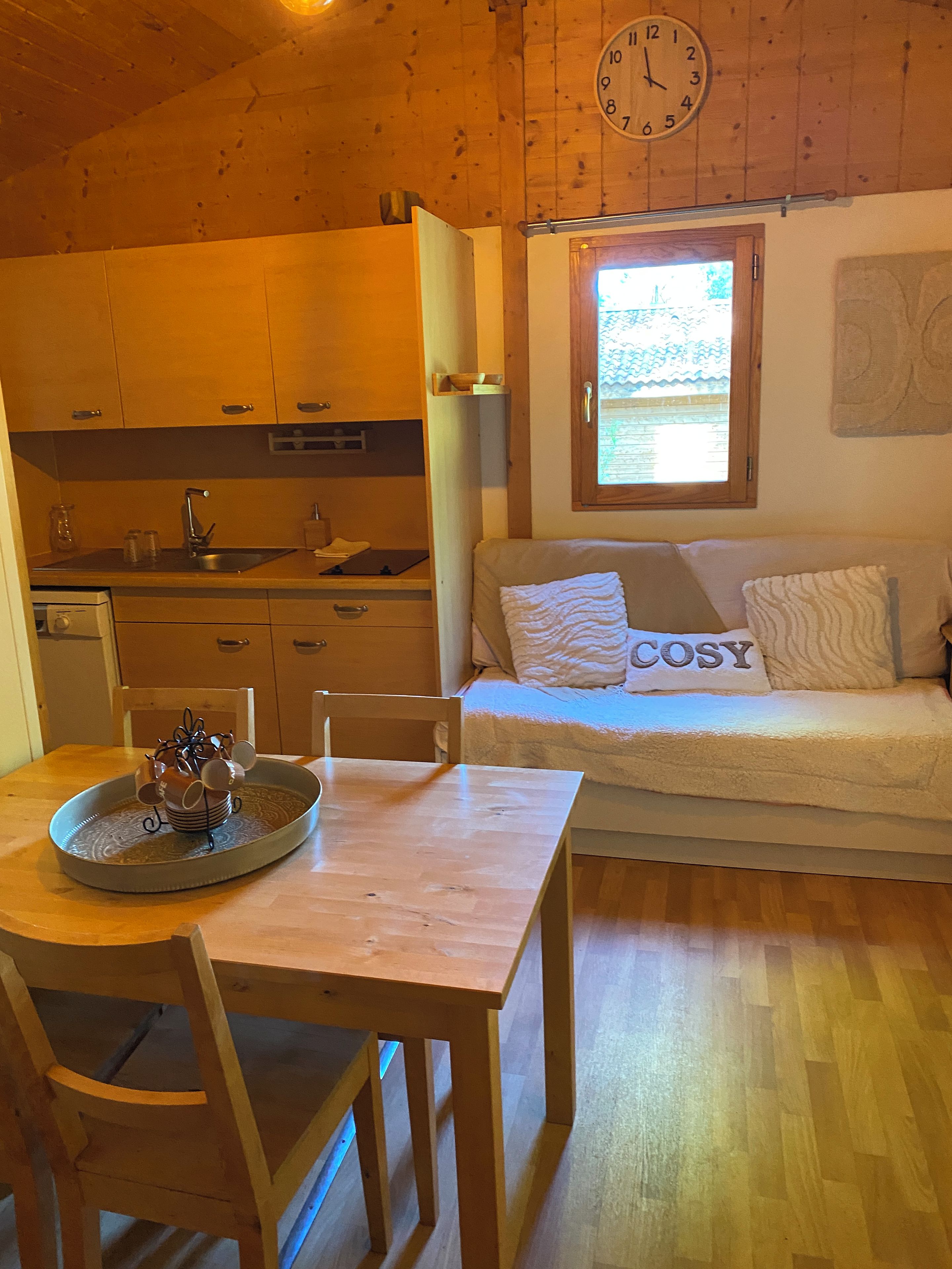 Een gezellig chalet interieur met een houten eettafel, een bank met een 'COSY' kussen, een keukenhoek en een raam met uitzicht.