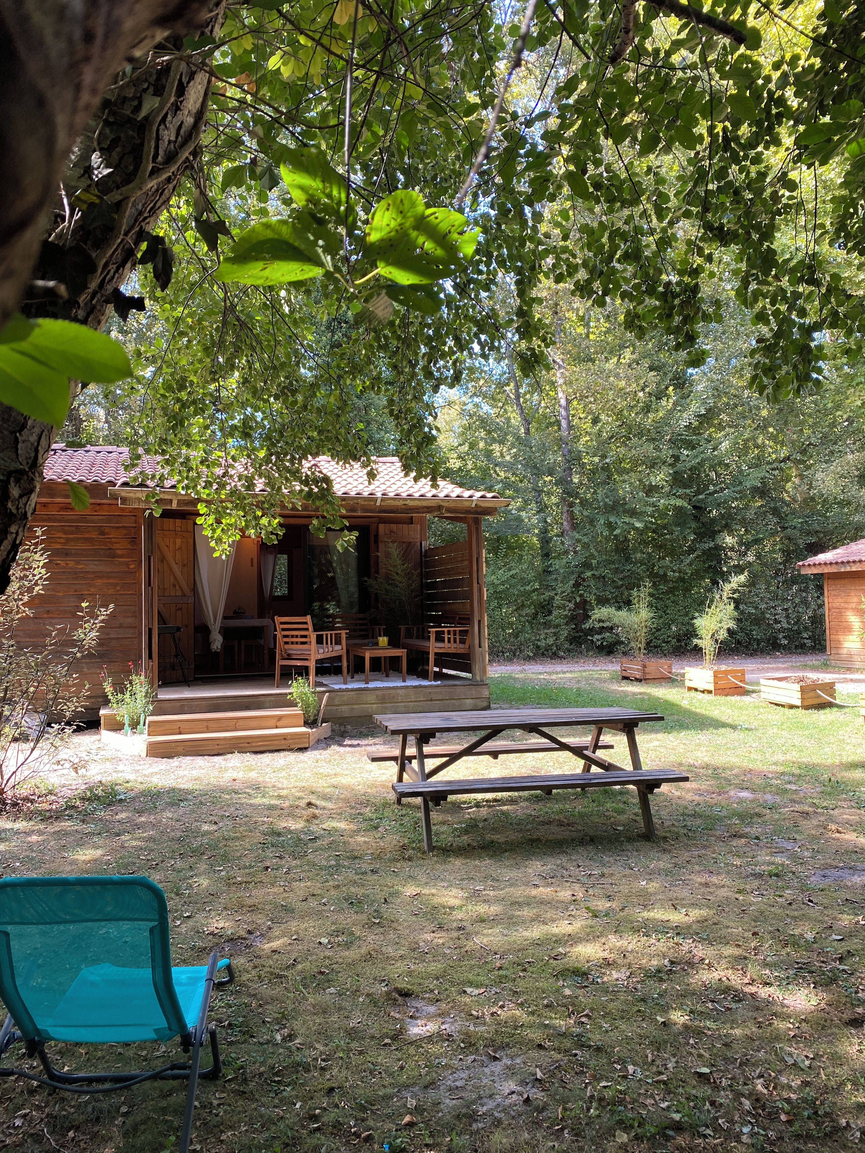 A wooden cabin with a porch, a picnic table, and a blue folding chair in a forested area.
