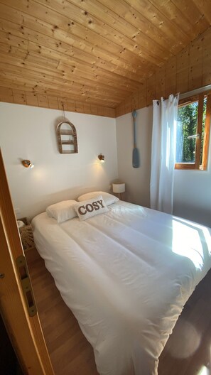 A bedroom with a wooden ceiling, a bed with white bedding, a wall-mounted mirror, and a window with curtains. - "Les Iris" Chalet, nestled in the heart of the Landes countryside, Casteljaloux (Pindères)