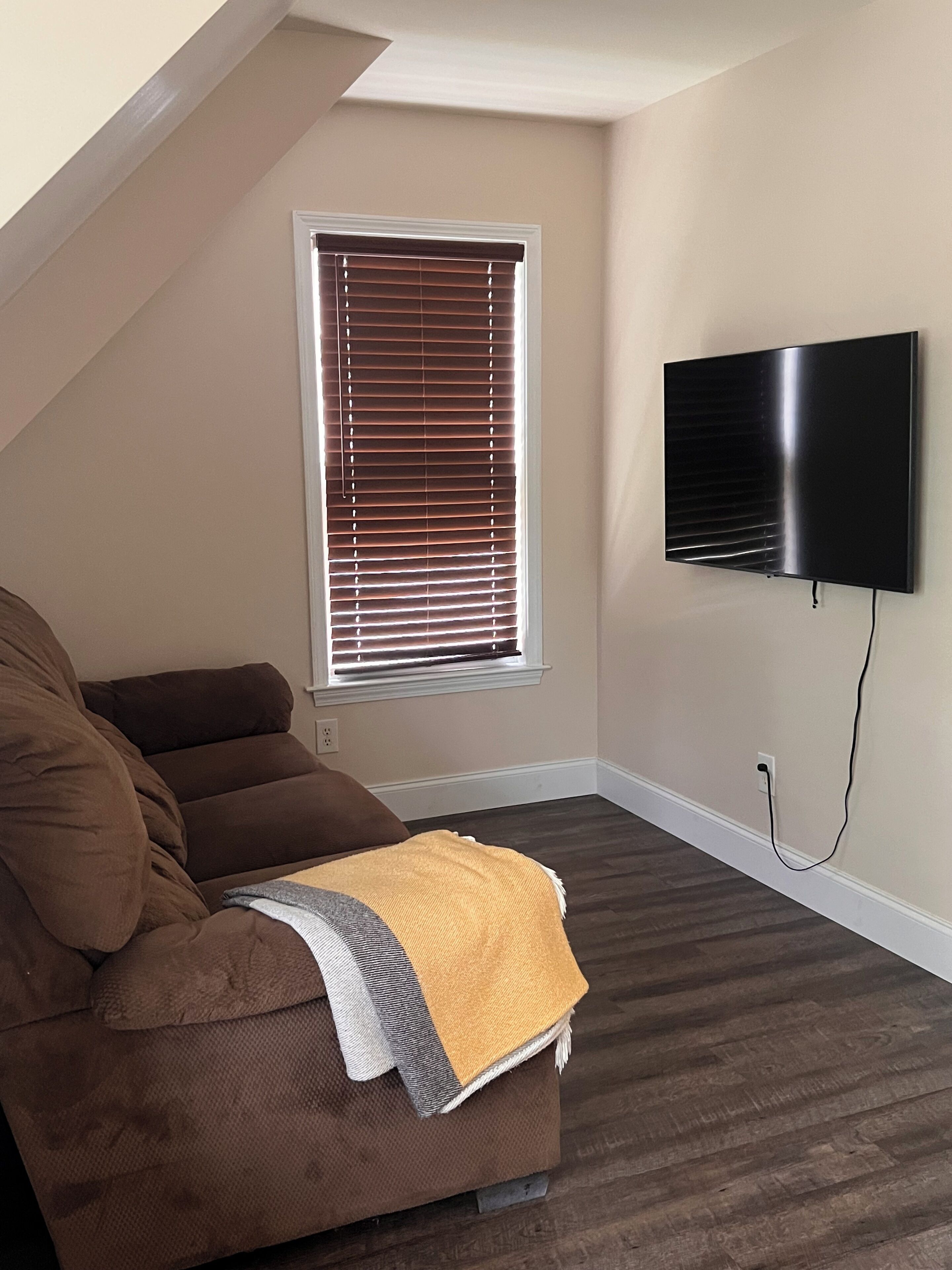 Ein Zimmer mit einer braunen Couch, einem an der Wand montierten Fernseher und einem Fenster mit Jalousien.