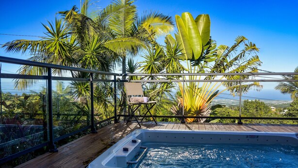Ein Whirlpool auf einer Holzterrasse mit Glastrennwand, mit Blick auf eine tropische Landschaft mit Palmen und Meerblick.