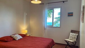 Ein Schlafzimmer mit Bett, einem Sessel, einem Fenster mit Blick auf Palmen und das Meer sowie einem Bild an der Wand.