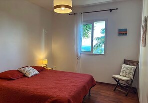 A bedroom with a bed, a chair, a window with a view of palm trees and the ocean, and a painting on the wall. - Ty Lodge Réunion – Pool, Spa, Ocean View, Garden, Near Saint-Pierre & Volcano (Le Tampon)