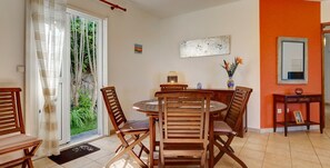 A dining area with a round wooden table and chairs, a small side table with a vase, and a door leading to a garden.