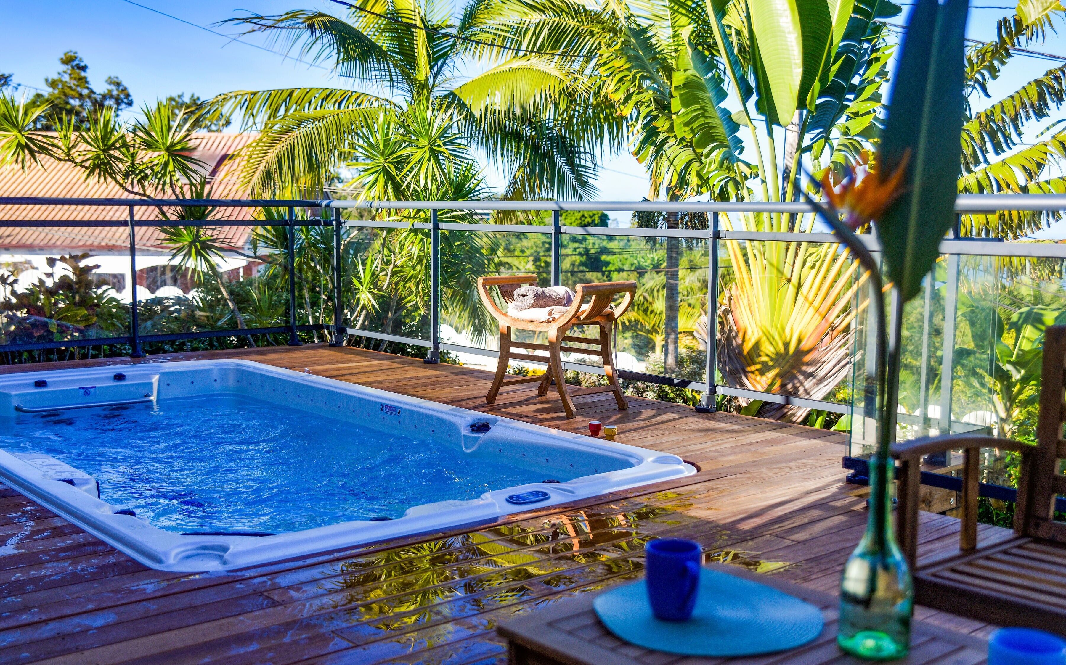 A hot tub on a wooden deck with chairs and a glass railing overlooking a tropical garden.