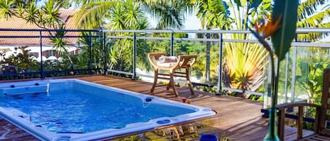 A hot tub on a wooden deck with chairs and a glass railing overlooking a tropical garden.