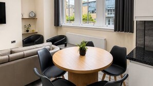 A modern living room with a round wooden table, black chairs, a beige sofa, and a large window with curtains. - Large Flat in Edinburgh 4/1 (Edinburgh)