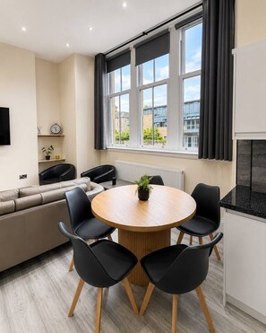 Ein modernes Wohnzimmer mit einem runden Holztisch, schwarzen Stühlen, einem beigen Sofa und einem großen Fenster mit Vorhängen.