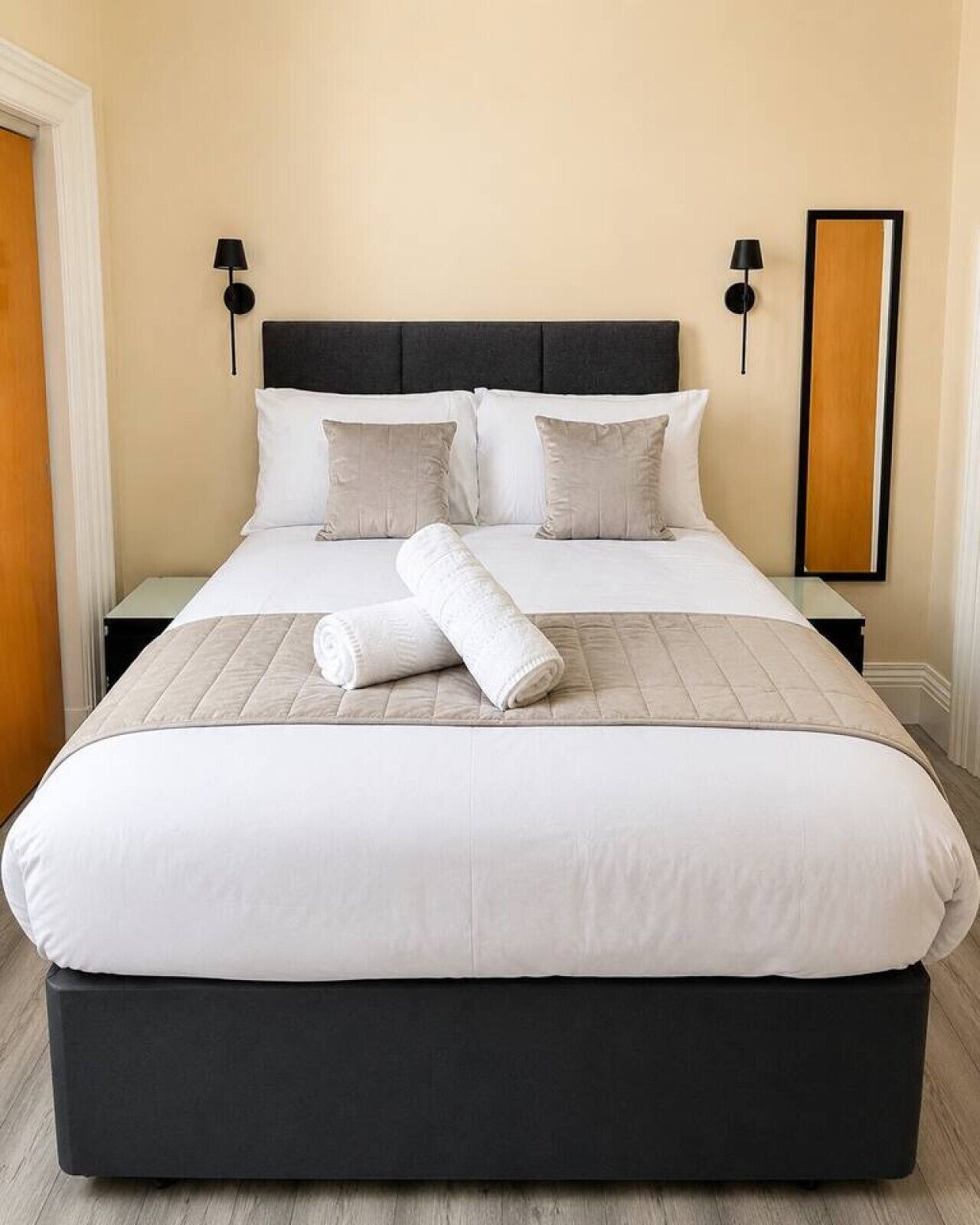 Een bed met een zwart hoofdeinde, wit beddengoed en twee bedlampjes.