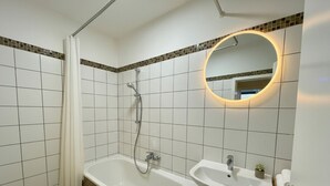 A bathroom with a bathtub, a sink, and a mirror with a circular light. - HomeBase with a kitchen and 5 single beds, centrally located (Krefeld)