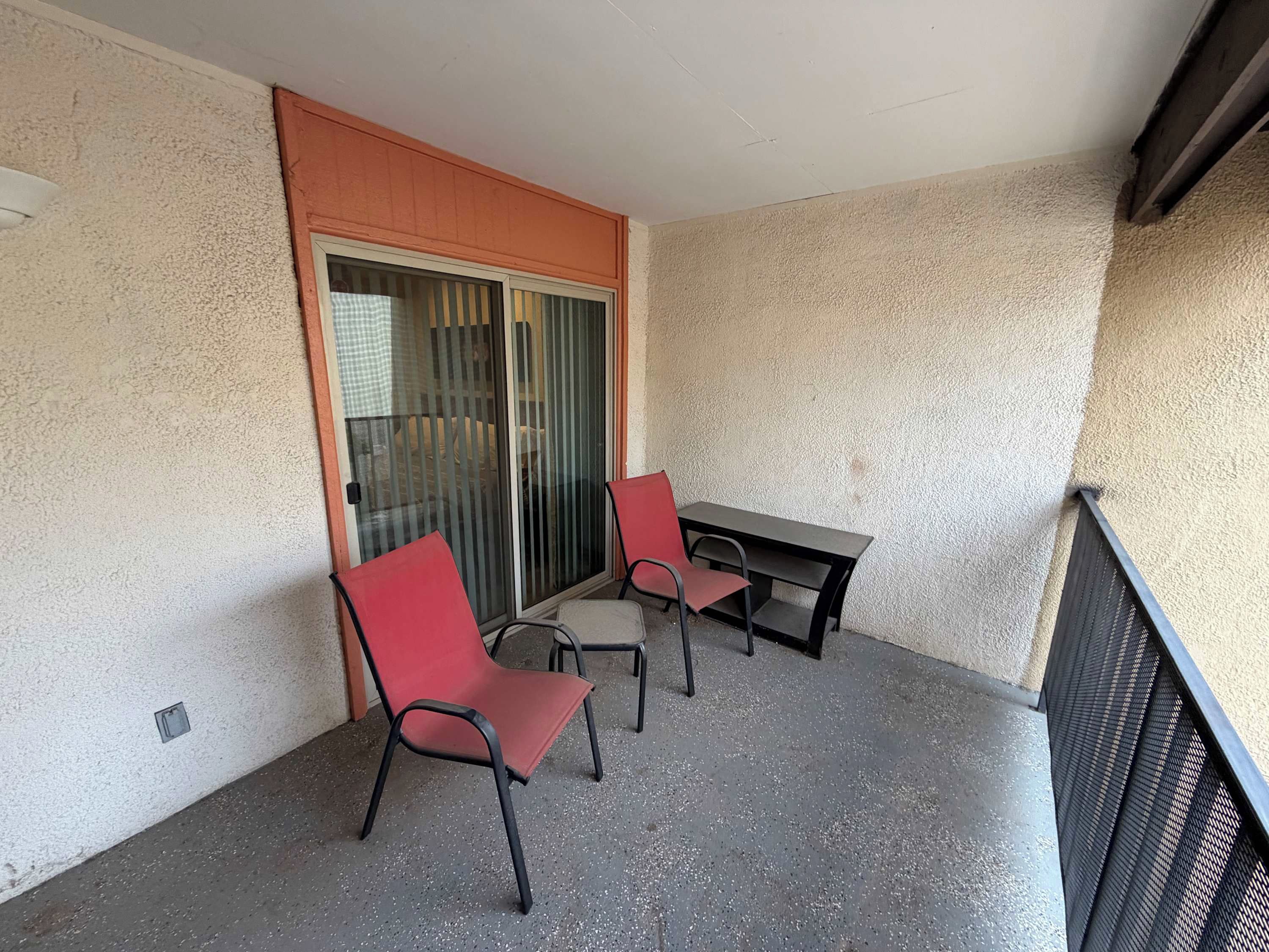 A balcony with two red chairs and a table, a sliding glass door, and a textured wall.