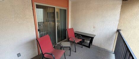 A balcony with two red chairs and a table, a sliding glass door, and a textured wall.