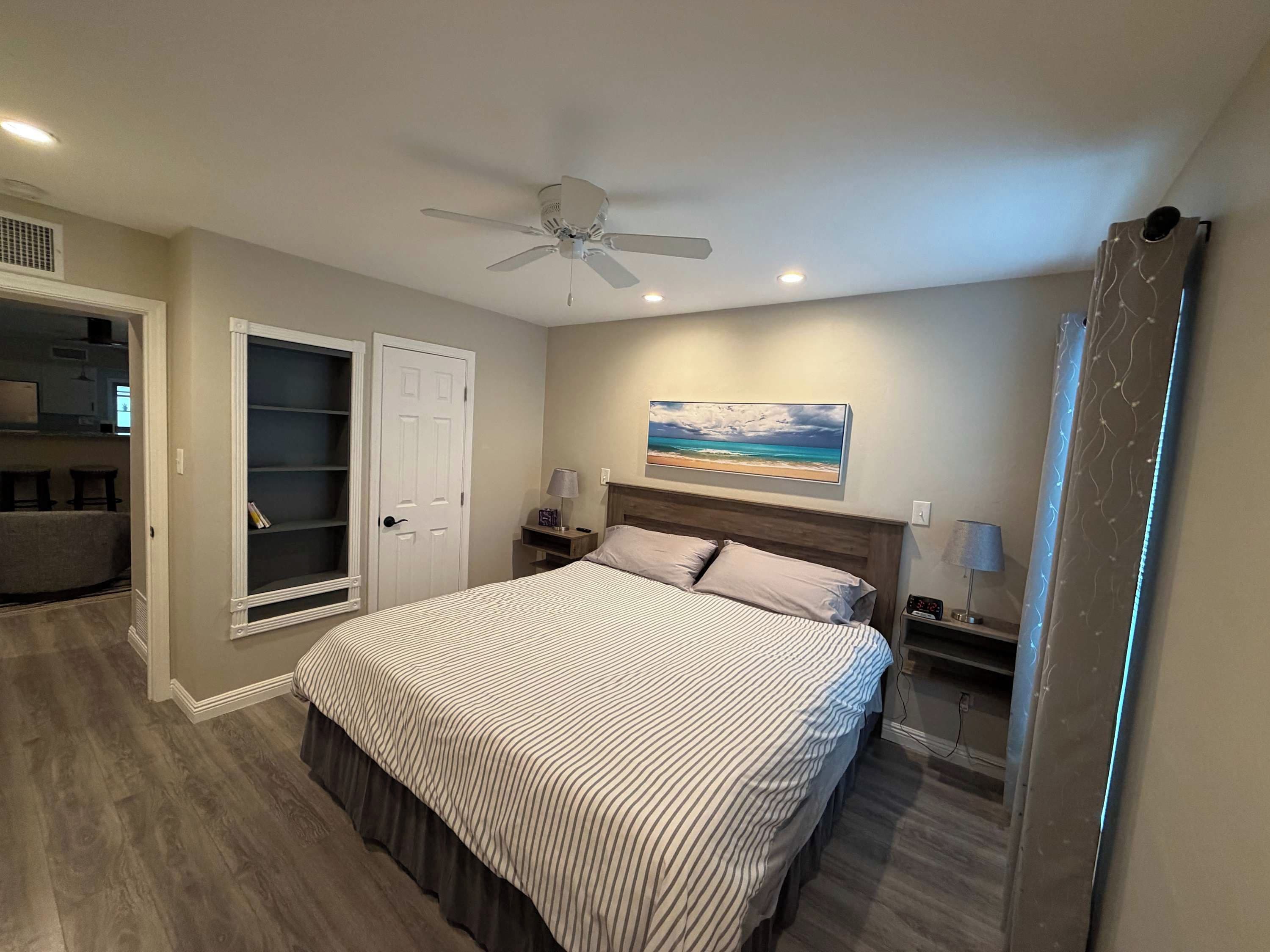 A bedroom with a bed, a ceiling fan, a nightstand, a lamp, a painting, and a wardrobe.
