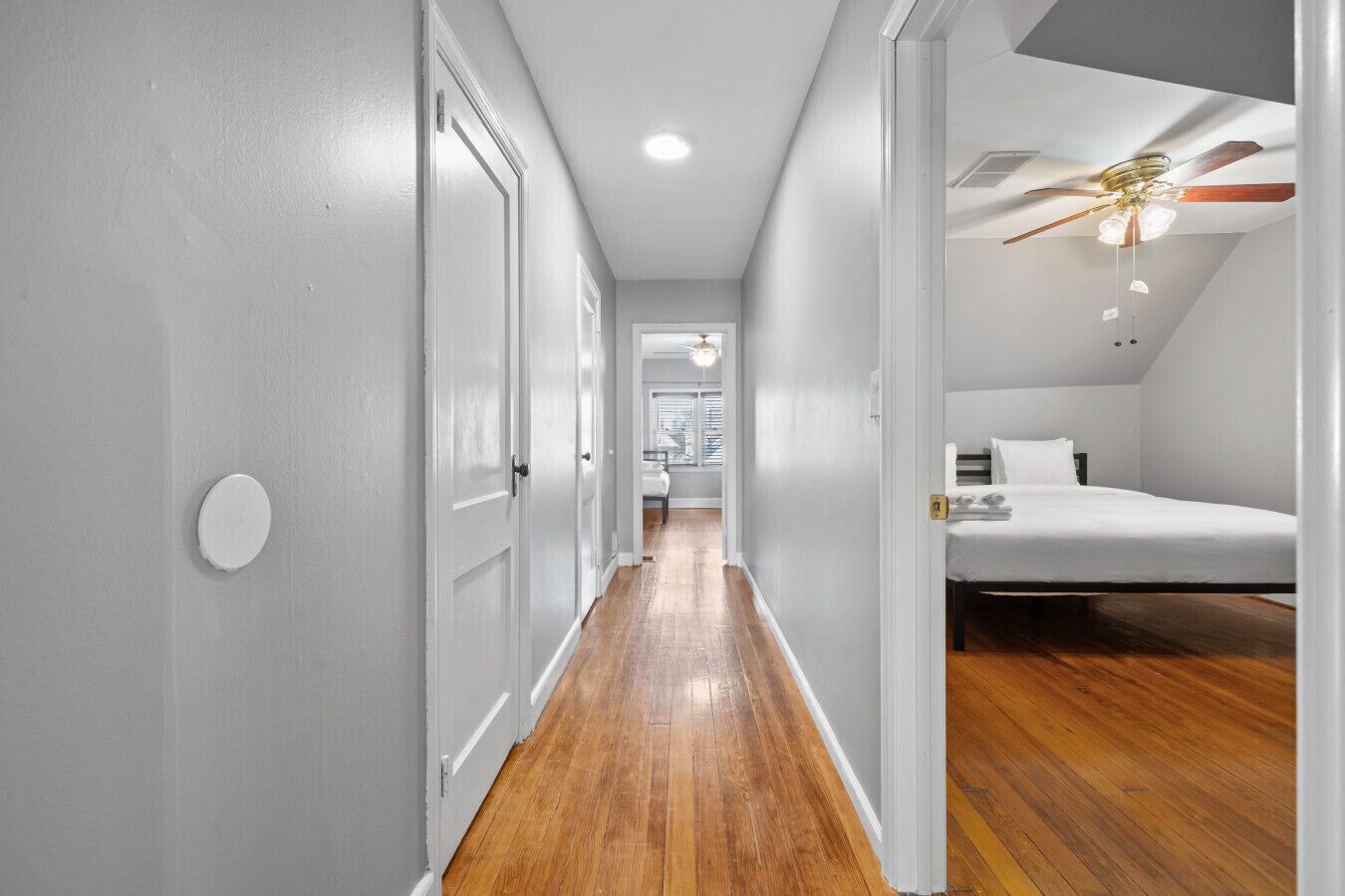 A narrow hallway with wooden flooring leading to a bedroom with a bed and a ceiling fan.