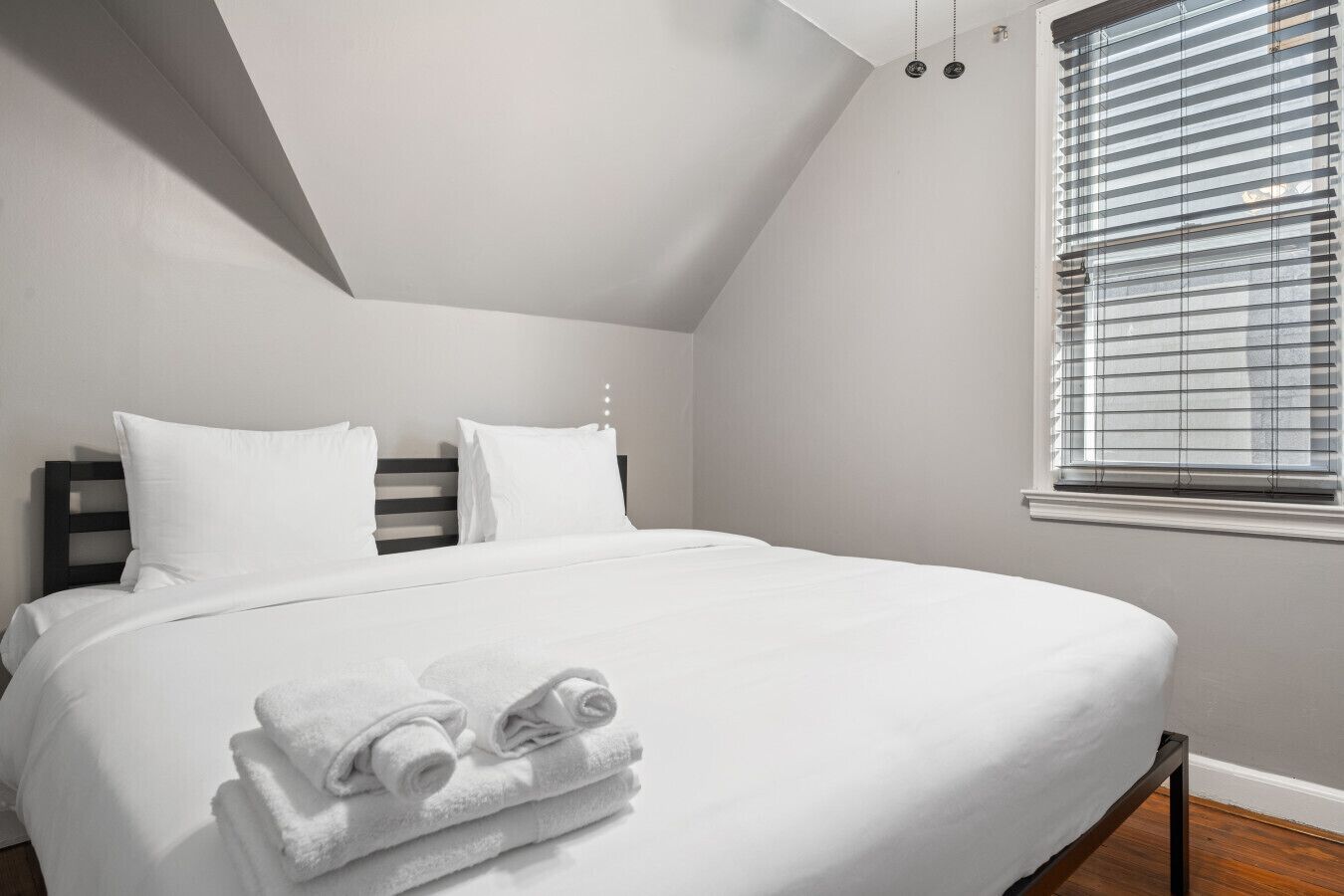 A neatly made bed with white linens and rolled towels, a dark headboard, and a window with blinds.