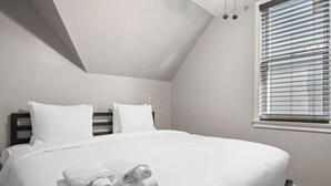 Una cama bien hecha con sábanas blancas y toallas enrolladas, un cabecero oscuro y una ventana con persianas.
