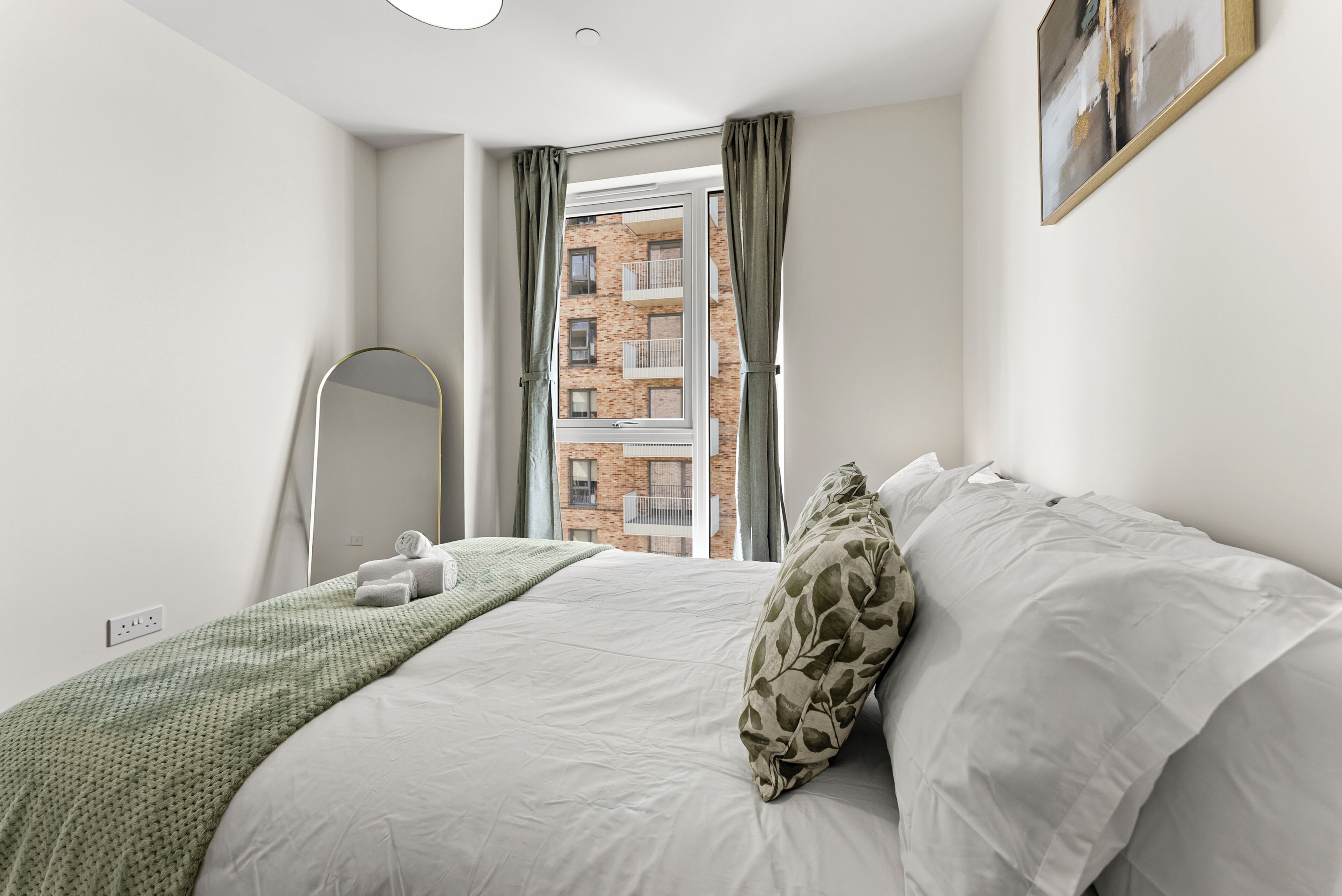 A bedroom with a bed, a mirror, a window with curtains, and a view of a building outside.