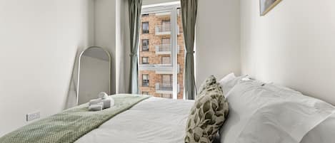 Ein Schlafzimmer mit einem Bett, einem Spiegel, einem Fenster mit Vorhängen und Blick auf ein Gebäude.
