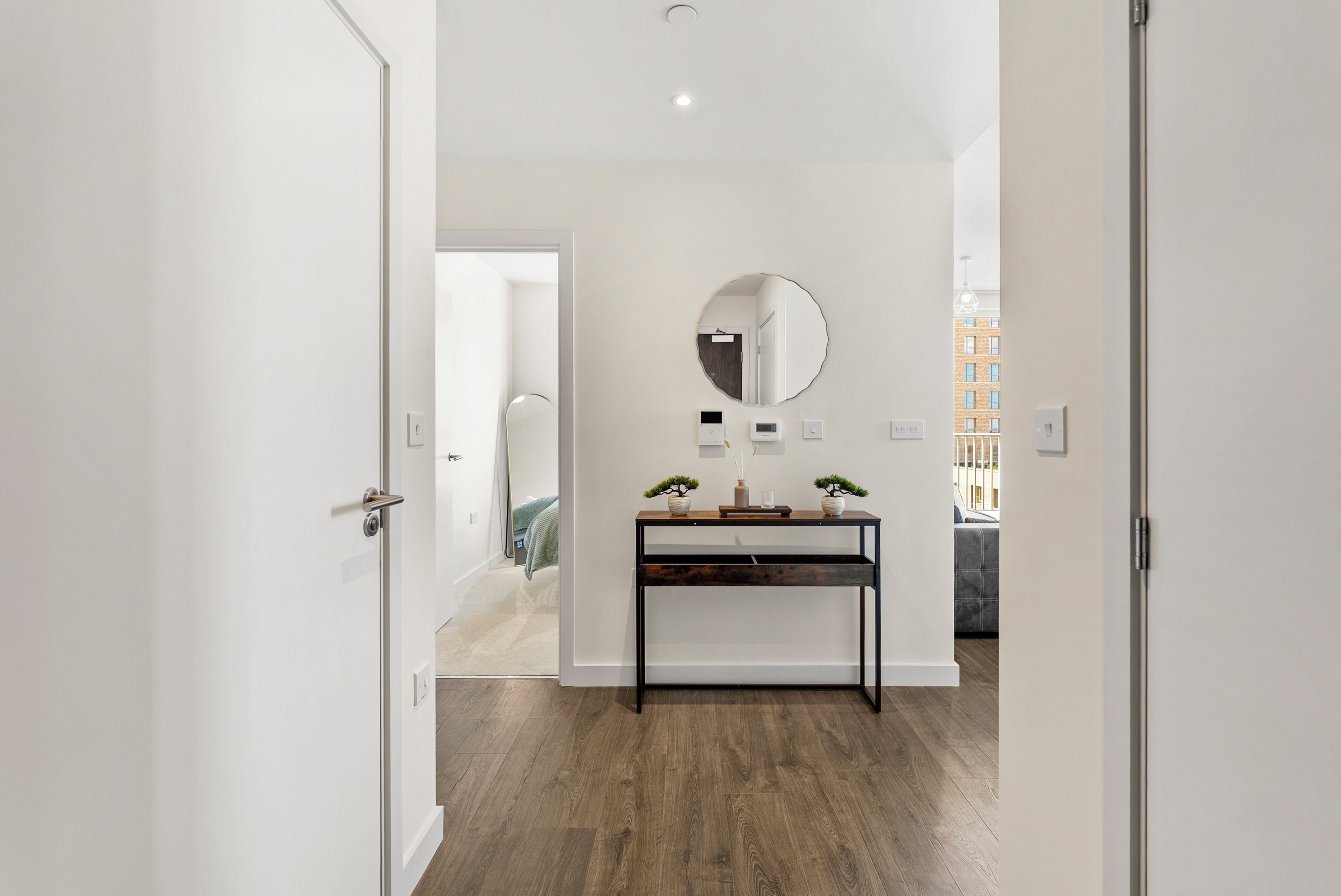 A hallway with a wooden floor, a black console table with two potted plants, a round mirror, and a door leading to another room.