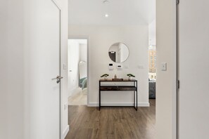 A hallway with a wooden floor, a black console table with two potted plants, a round mirror, and a door leading to another room. - Stylish 2 BR Apt | Sleeps 8 | Balcony | 2BA | Near Wembley (Harrow)