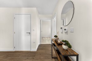 Un pasillo con piso de madera, paredes blancas, un banco de madera con un florero y plantas, y un espejo redondo en la pared.