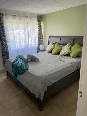 A hotel room with a bed, curtains, a bedside table, and a nightstand. - Fresher Breeze, your home from home in the heart of the beautiful Ocho Rios (St Anns)