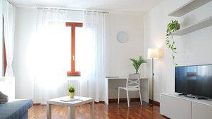 A modern living room with a sofa, a coffee table with a plant, a desk with a chair, a television, and a clock on the wall. - Double Space Apartment - Milan - 6 guests · Airport · Train · Garage (Milano)