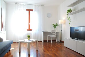 Ein modernes Wohnzimmer mit einer Couch, einem Couchtisch mit Pflanze, einem Schreibtisch mit Stuhl, einem Fernseher und einer Uhr an der Wand.