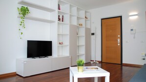 A modern living room with a flat-screen TV, white shelving units, a small white table with a plant, and a wooden door. - Double Space Apartment - Milan - 6 guests · Airport · Train · Garage (Milano)