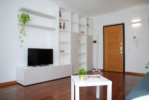 Ein modernes Wohnzimmer mit einem Flachbildfernseher, weißen Regalelementen, einem kleinen weißen Tisch mit einer Pflanze und einer Holztür.