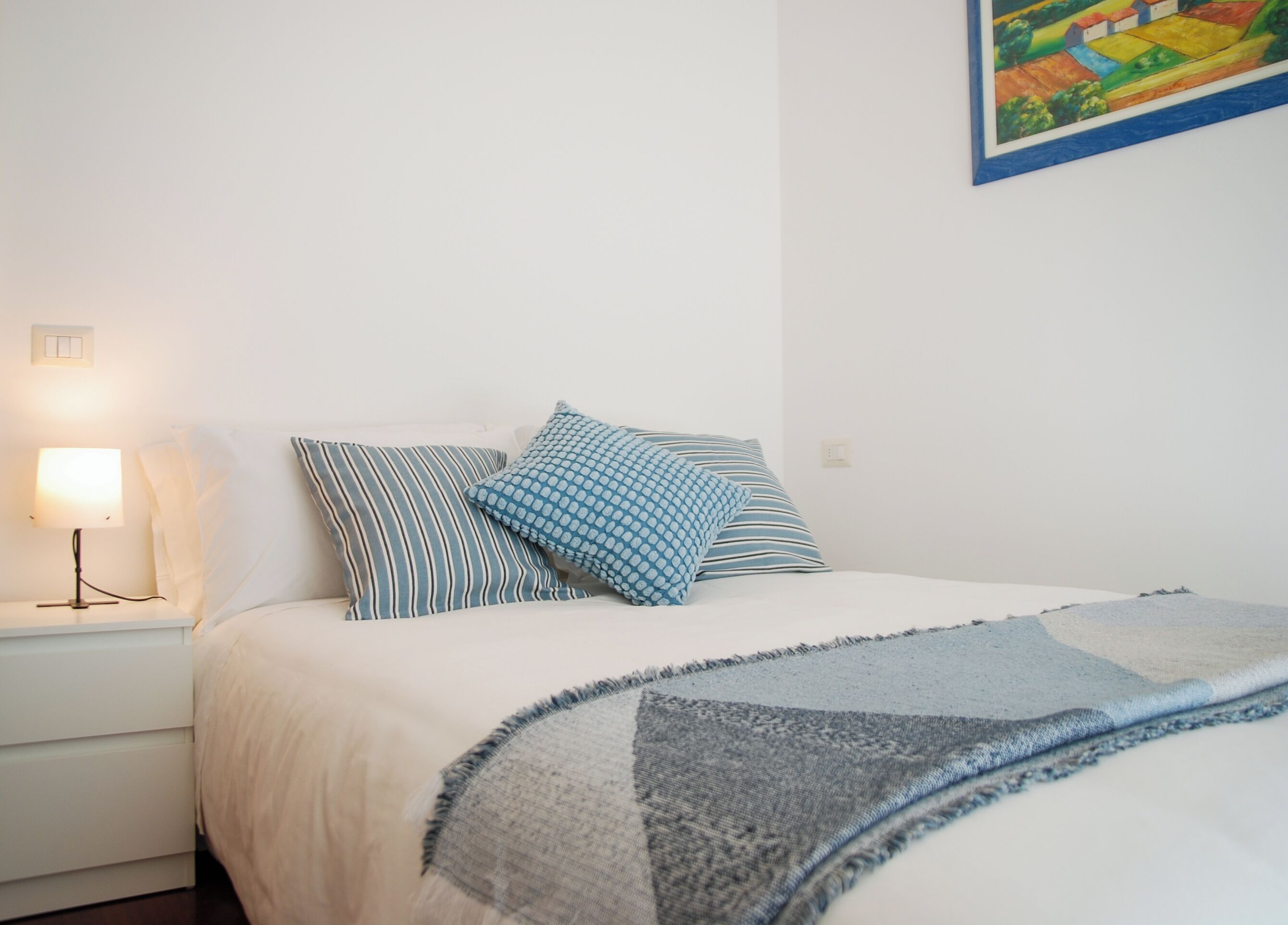 A bedroom with a bed, pillows, a bedside lamp, and a painting on the wall.