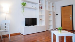 A living room with a flat-screen TV on a white entertainment unit, a white chair, a small table with a plant, and a wooden floor. - Double Space Apartment - Milan - 6 guests · Airport · Train · Garage (Milano)