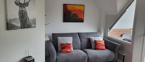 A modern living room with a grey sofa, two cushions, a colorful rug, a painting of a deer, and a window with a view of a brick wall.