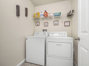 Une buanderie équipée de deux machines à laver et deux sèche-linge superposés, d’une étagère contenant des produits de nettoyage et de tableaux encadrés accrochés au mur.