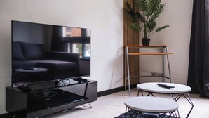 Ett modernt vardagsrum med en platt-TV, en soffa, ett soffbord och ett litet sidobord. Det står en krukväxt på ett träställ och det finns ett fönster med utsikt.