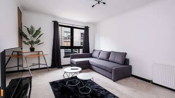 A modern living room with a grey sofa, a small white table, a black rug, a potted plant, and a ceiling light fixture.